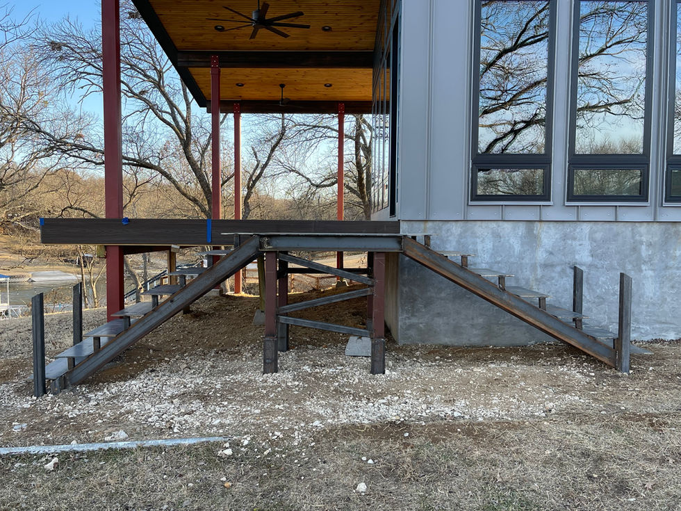 A partially constructed outdoor staircase with custom fabrication, featuring metal framing and wooden steps, leads up to a modern house with large windows and a covered porch. The ground is bare and scattered with gravel and dirt.