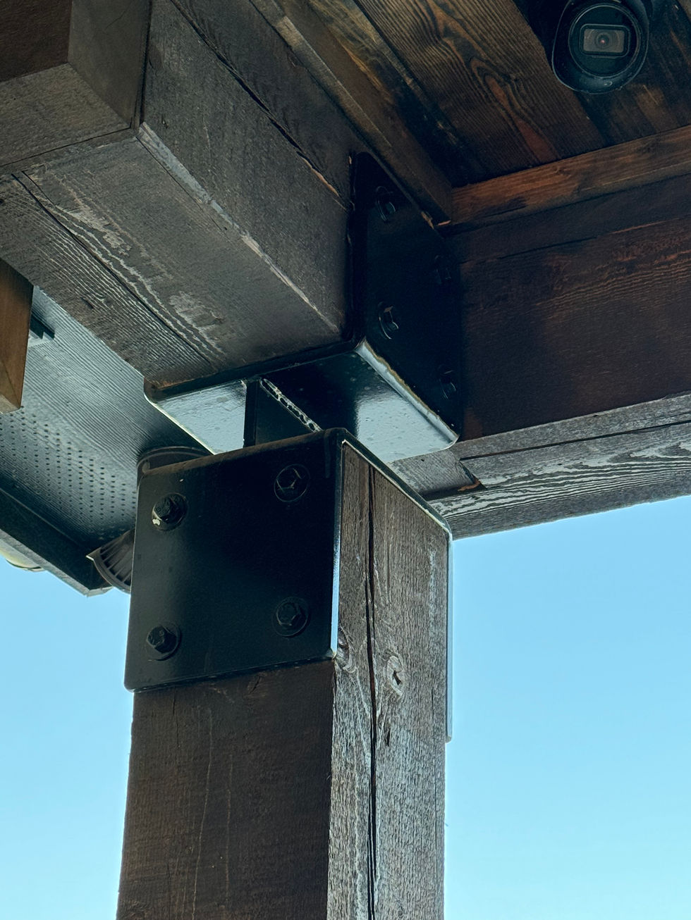 Close-up view of a wooden beam structure connected with industrial style brackets and bolts, set against a clear blue sky; custom fabrication is evident, and a security camera is visible in the upper right corner.