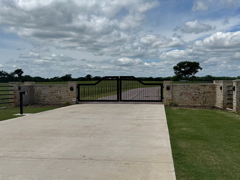 A black metal custom gate set in a stone wall blocks the entrance to a driveway, with green grass and open fields in the background under a partly cloudy sky, showcasing expert custom fabrication.