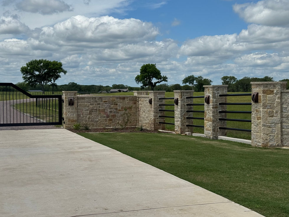 A stone and metal fence with a closed black custom gate borders a grassy yard and extends along a rural road under a partly cloudy sky, with trees and open fields in the background.
