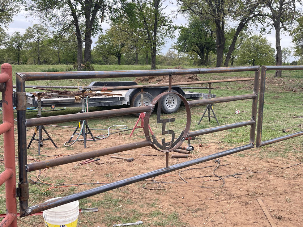 Two large metal sculptures featuring the logo L5 inside circles sit on a workbench in an industrial workshop, showcasing custom fabrication skills with red welding screens, tools, and a view of a grassy field through an open door.