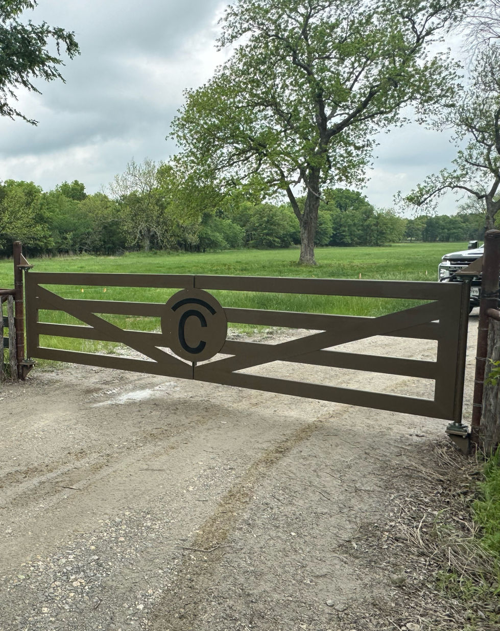 A modernized steel gate with a C emblem in the center stands across a dirt driveway, showcasing custom gates design, with green grass, trees, and a parked vehicle visible in the background on a cloudy day.