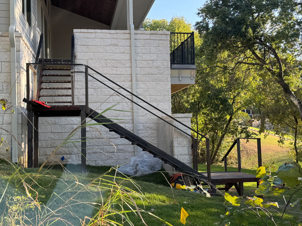 A modern house with white stone walls features a custom staircase of black metal leading from the deck to the grassy backyard. Trees surround the area, and some construction materials hint at ongoing residential construction near the stairs.