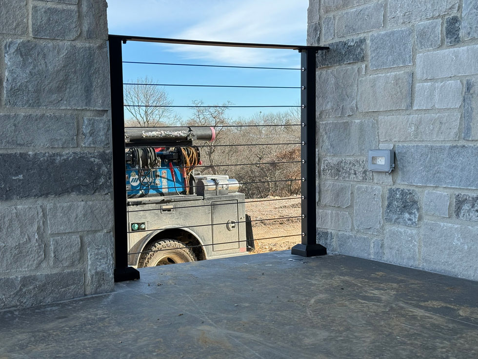 A custom railing in sleek black metal is installed between stone walls on a patio; outside, a utility truck with equipment on its bed is parked on a dirt area with trees and blue sky in the background.