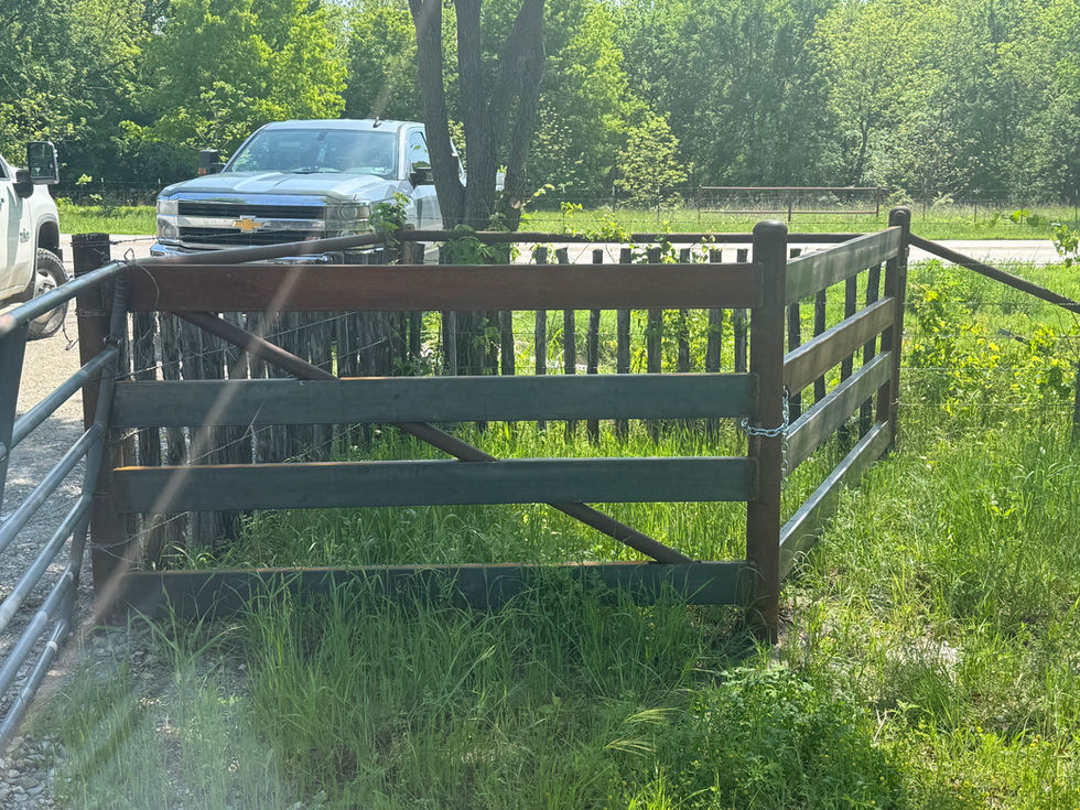 A small, metal trash can enclosure with a chained gate stands in a grassy area near trees. Two white pickup trucks are parked nearby, with one person possibly performing mobile welding by equipment in the background.