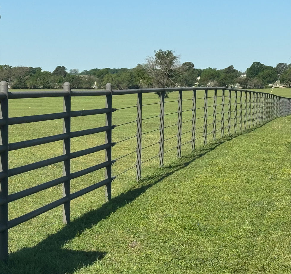 A long black cable fence stretches diagonally across a grassy field under a clear blue sky, with trees and scattered buildings in the background.