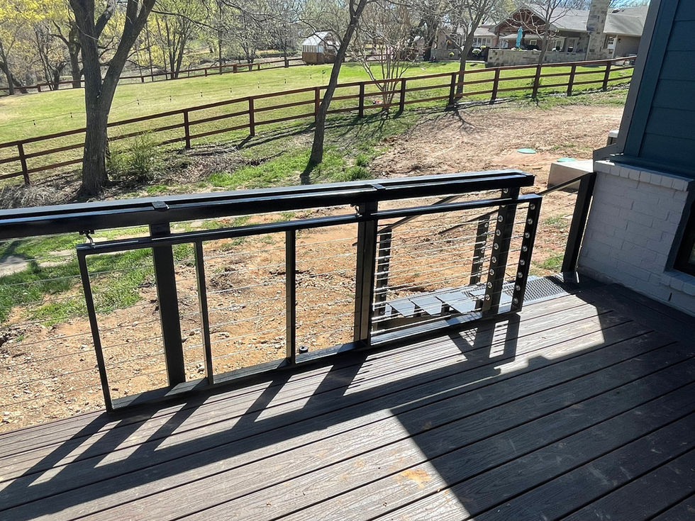A modern house with a covered deck featuring custom railing and cable wires overlooks a body of water. The exterior showcases blue and white siding, while outdoor stairs lead down to a gravel and dirt yard, highlighting quality residential construction.
