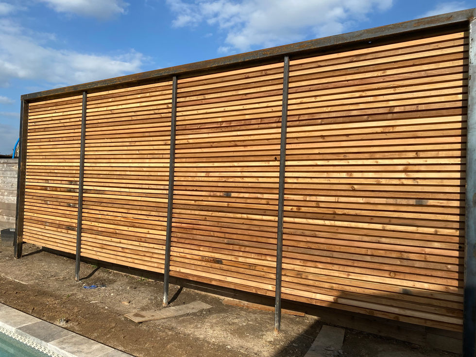 A large wooden privacy wall with horizontal slats, supported by vertical metal beams likely installed through mobile welding, stands beside a patch of bare dirt under a partly cloudy sky.