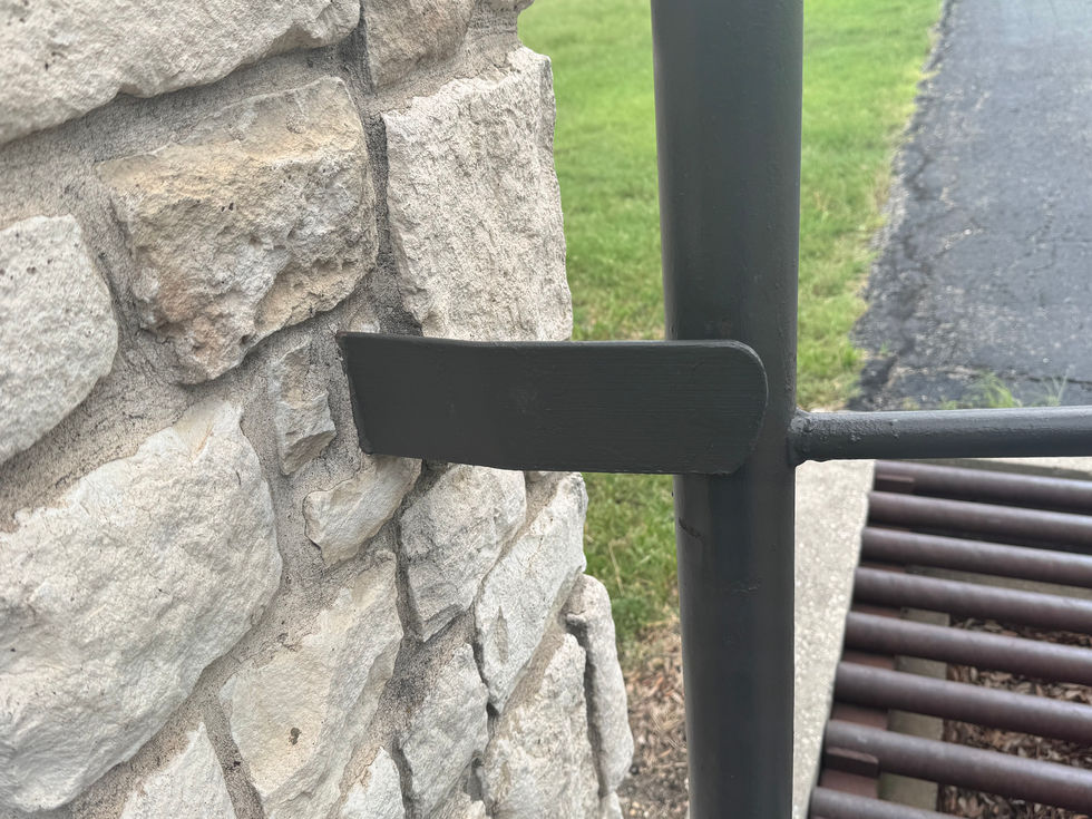 A close-up of a black metal latch on a contemporary gate rests against a light-colored stone wall, with grass and part of an asphalt path visible in the background.
