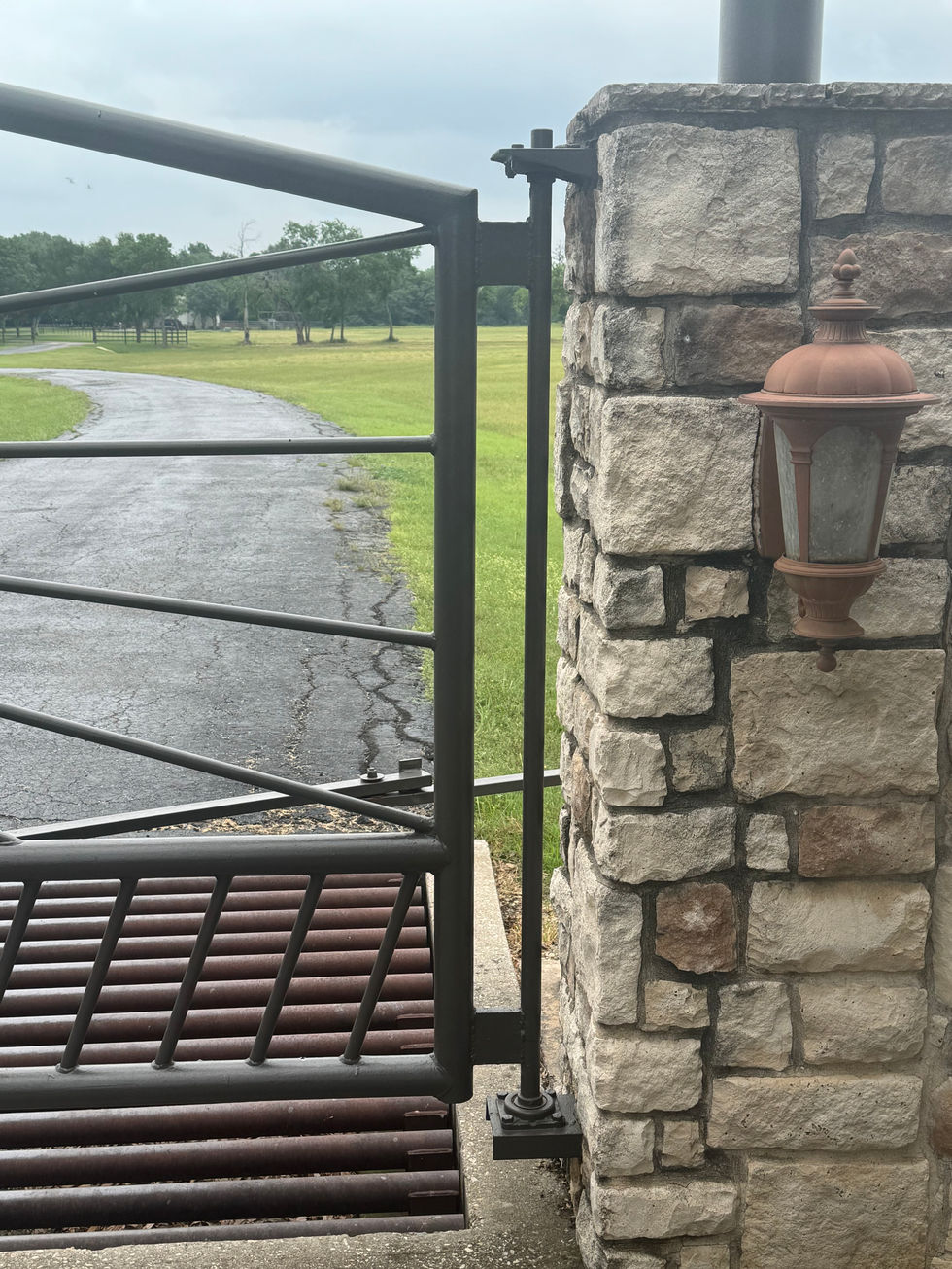 A custom gate attached to a stone pillar stands open next to a paved driveway and grassy field. A brown outdoor lantern is mounted on the pillar, highlighting the custom fabrication against the overcast sky.