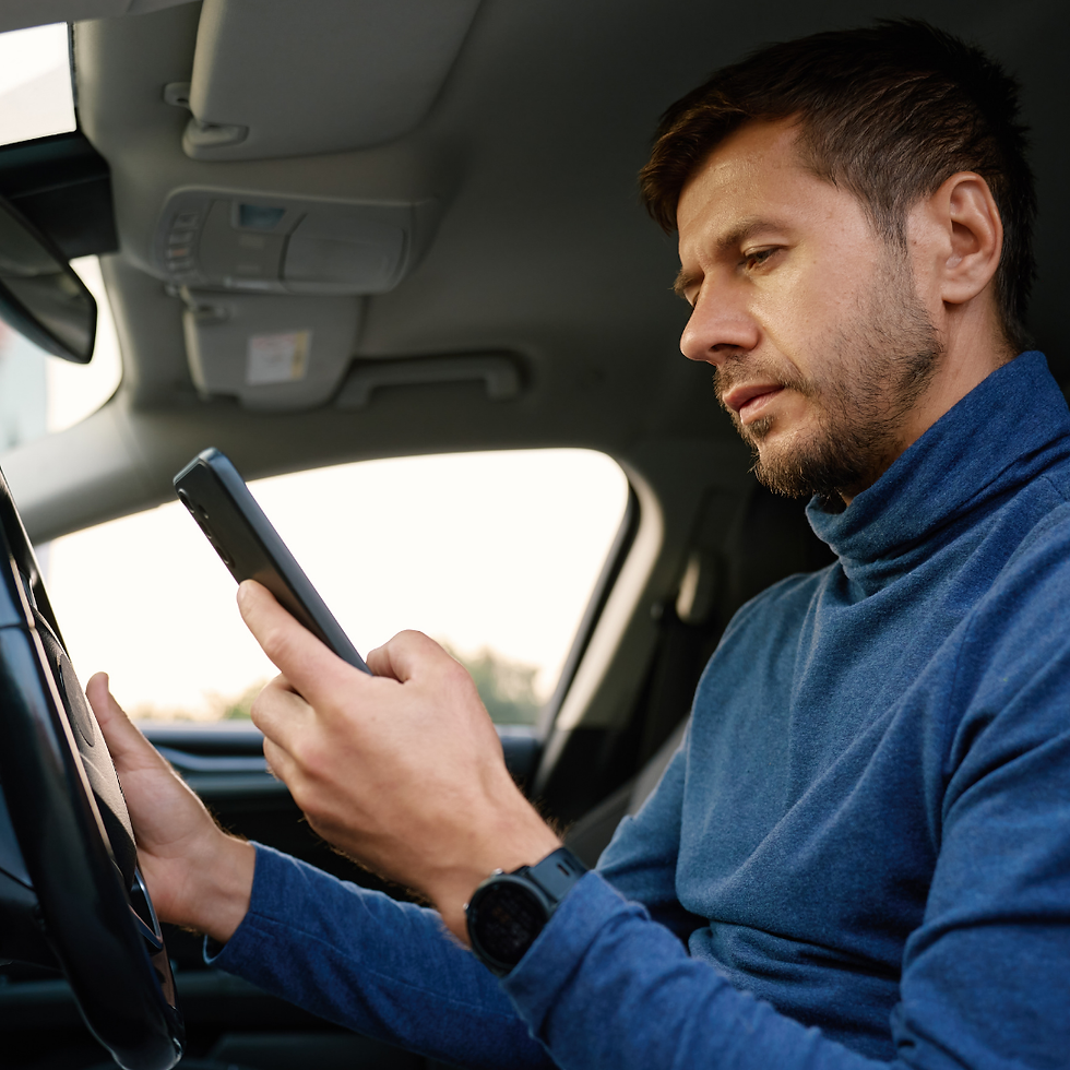 Man in a blue turtleneck sits in a car, looking at a smartphone with a focused, slightly serious expression.