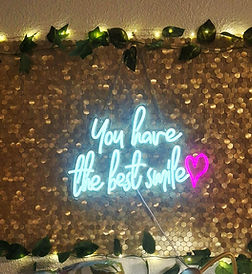 Gold sequin backdrop with neon sign 'You have the best smile' and green leaves.