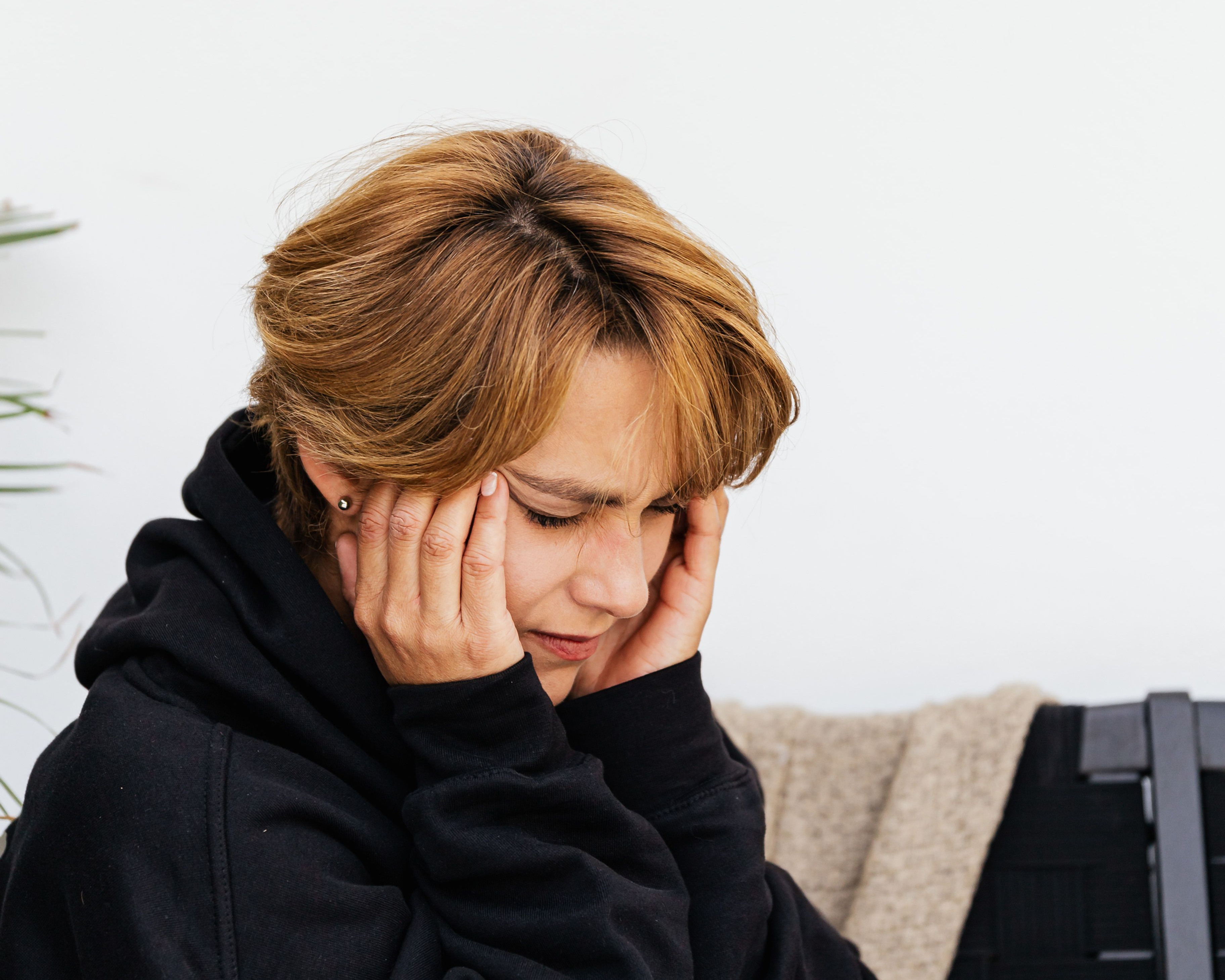 Distressed woman in a black hoodie and ripped jeans holds her head while sitting indoors against a white wall.