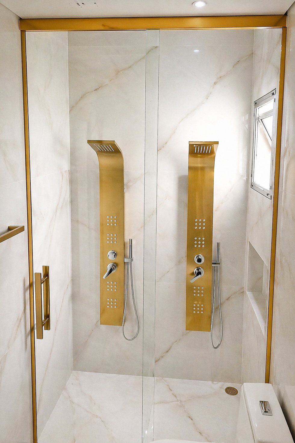 Duas colunas de chuveiro douradas em um box de vidro com acabamento dourado. Parede de mármore, janela pequena à direita. Ambiente luxuoso e moderno.