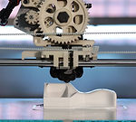 Figura blanca orgánica sobre una mesa, siendo impresa por una impresora 3d metalica con diferentes engranes 