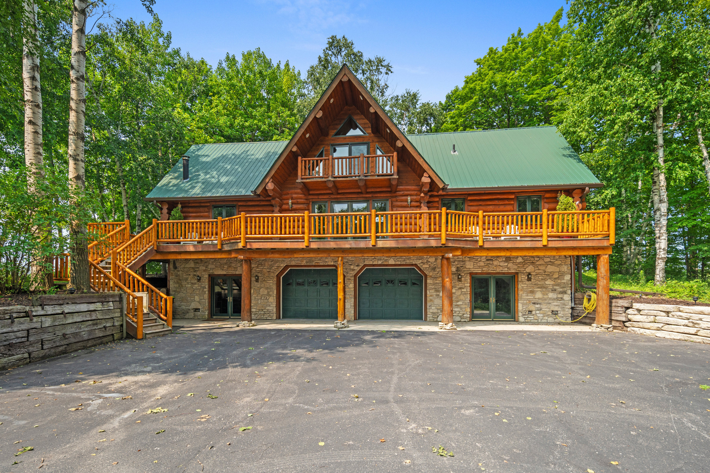 Two-story log cabin with a green roof and wraparound deck among tall trees