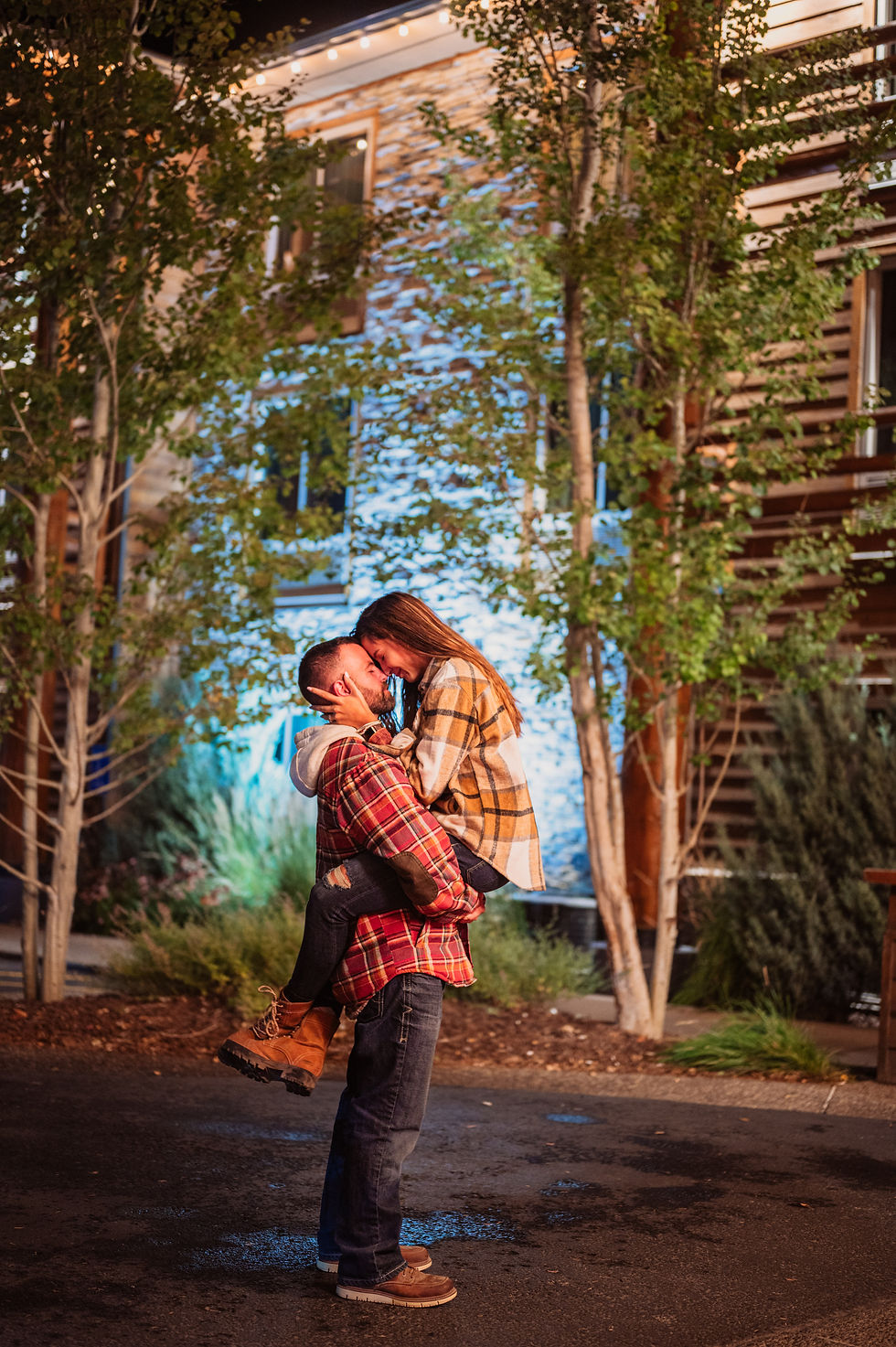 A couple embraces joyfully at night; he lifts her as they smile. They're outdoors, surrounded by trees and a lit building in warm plaid.