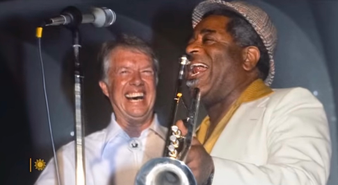 two men having a great time on stage during a music event. they're standing in front of the microphone as one of the men plays a saxaphone