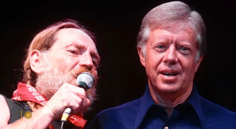 Two middle aged white men appear to be on stage at music event. Man on the left singing in microphone is Willie Nelson standing next to Jimmy Carter about 30 years ago. 