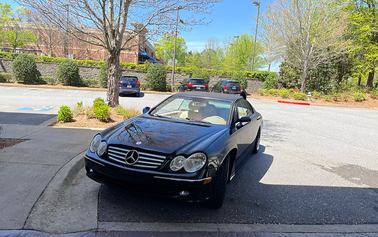 Front view of a black Mercedes-Benz in a concrete parking lot with cars, trees, and a brick building in the background, featured in Tolon Detail Services' luxury auto detailing showcase.