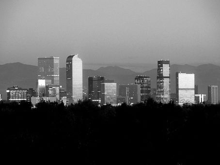 Black and white photo of city skyline at dusk, with illuminated skyscrapers and mountains in the background, creating a serene mood.
