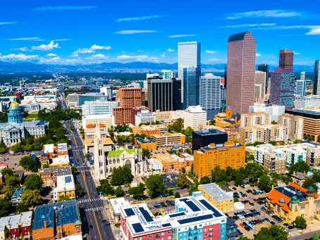 Aerial view of a cityscape with skyscrapers, historical buildings, and greenery against a backdrop of mountains under a blue sky.