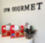 IFM GOURMET sign above festive Christmas gift bags and a pine-cone floral arrangement with red berries on a white shelf.
