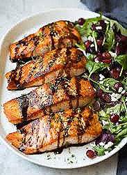 Grilled salmon fillets drizzled with sauce on a white plate beside a salad with greens, red grapes, and feta on a marble surface.
