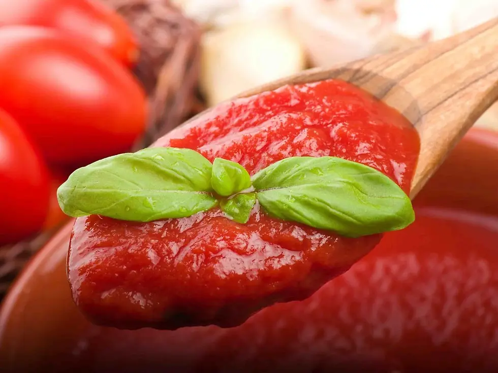 Wooden spoon lifts glossy tomato sauce topped with basil, with blurred tomatoes in the background.