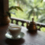 White teacup on saucer with tea, next to a bronze teapot on a wooden table. Lush green foliage visible through a window, serene mood.