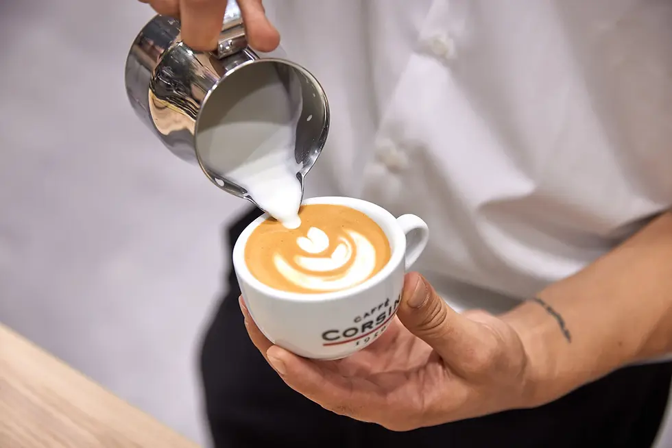 Barista pouring steamed milk into a cappuccino cup forming latte art. The cup has logo text, and there's a white shirt in the background.
