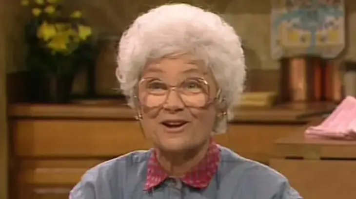 Image of Sophia Petrillo from The Golden Girls, wearing large glasses, a denim shirt with a pink collar, and smiling brightly in a kitchen setting.