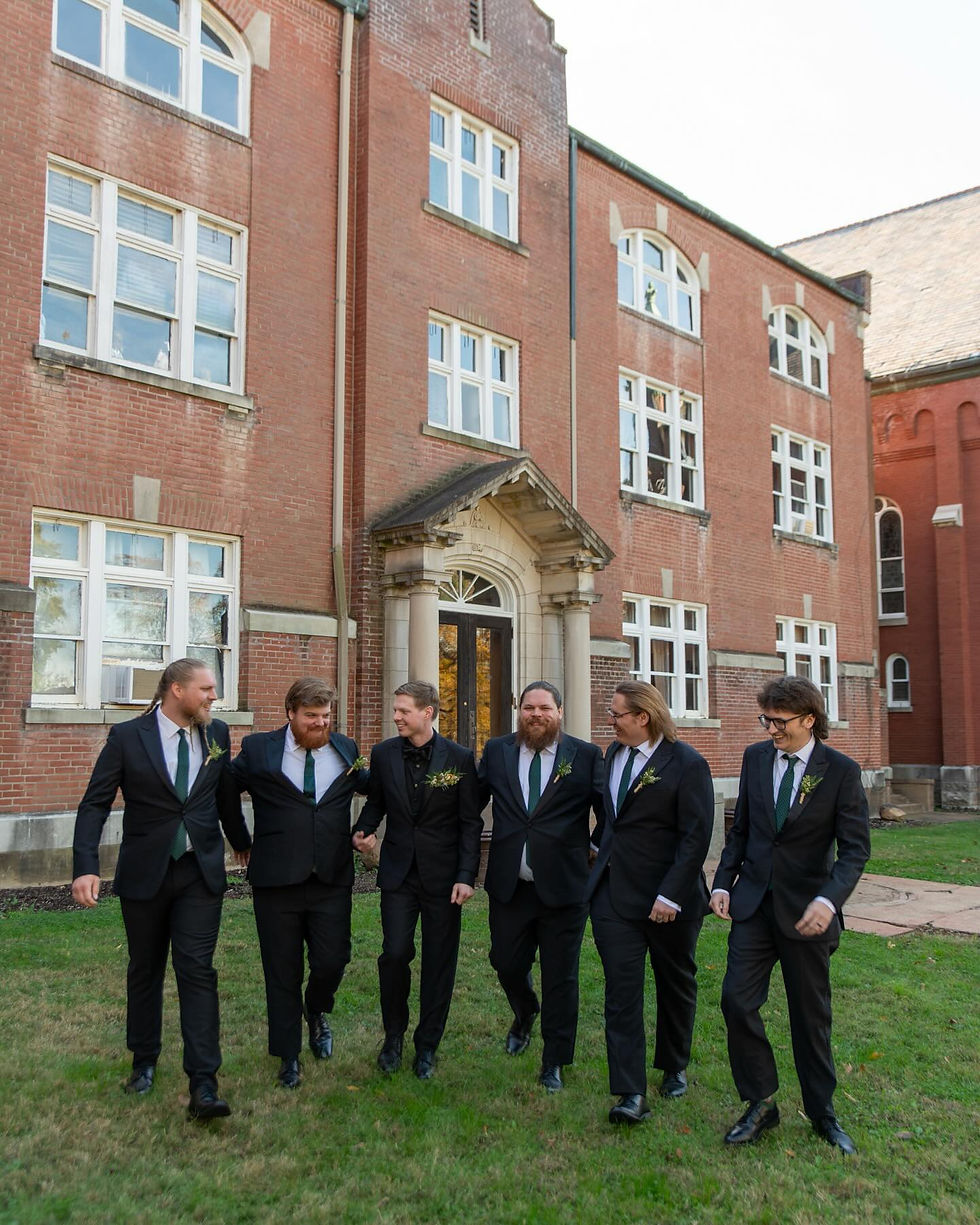 Groomsmen in black suits walk arm-in-arm, smiling, in front of a arcadia academy with white windows, on a grassy lawn.
