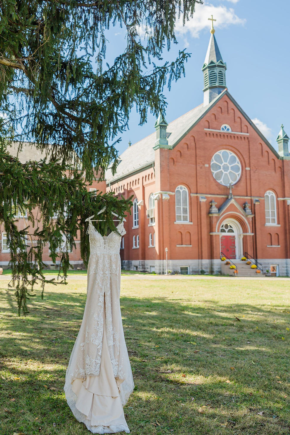 Wedding dress hangs from a pine tree in front of a red brick church with steeple under a bright blue sky.