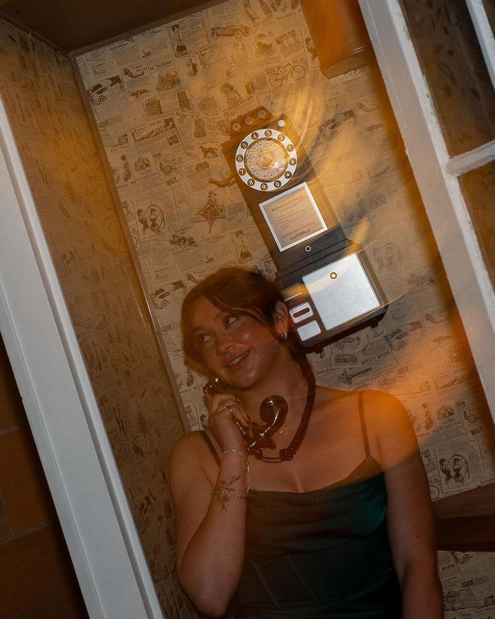 Woman in a dark dress smiles while using a vintage rotary phone in a phone booth with newspaper-patterned wallpaper. Warm lighting.