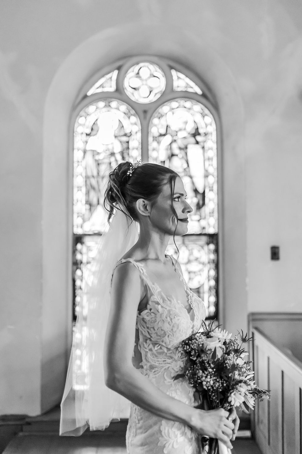 Bride in lace gown and veil holds bouquet in a church before stained-glass windows, black-and-white, serene profile view
