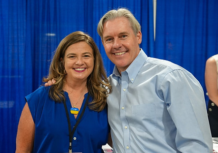 Lee Johnson poses for a picture with a lady wearing blue in front of a blue curtain.