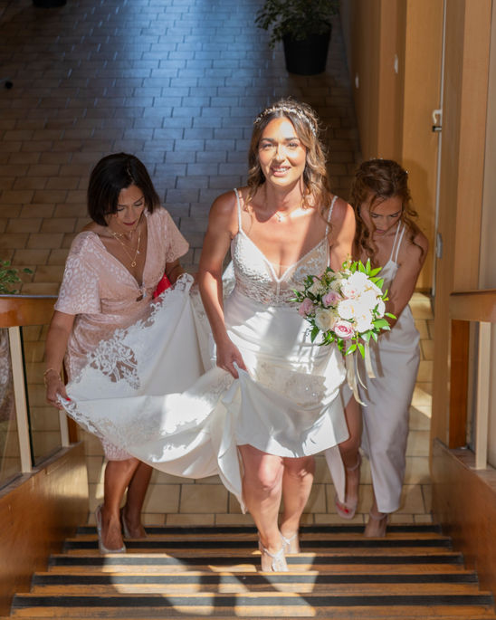 Photographie de mariage en Île-de-France, la mariée monte l'escalier à la mairie de Jouy-le-Moutier avec une magnifique robe blanche qui tiennent deux dames d'honneur et sourie avec bonheur.