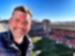 Sports travel Tom, Smiling man in a black jacket takes a selfie at a lively soccer stadium under a clear blue sky.