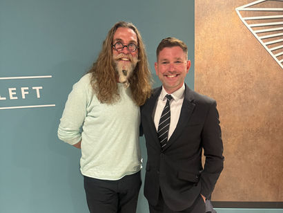 Two men stand smiling in front of a blue wall with "LEFT" on it. One wears glasses and a light shirt, the other a black suit and tie.