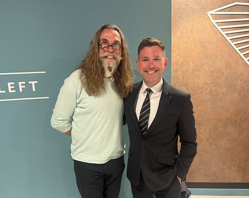 Two men stand smiling in front of a blue wall with "LEFT" on it. One wears glasses and a light shirt, the other a black suit and tie.