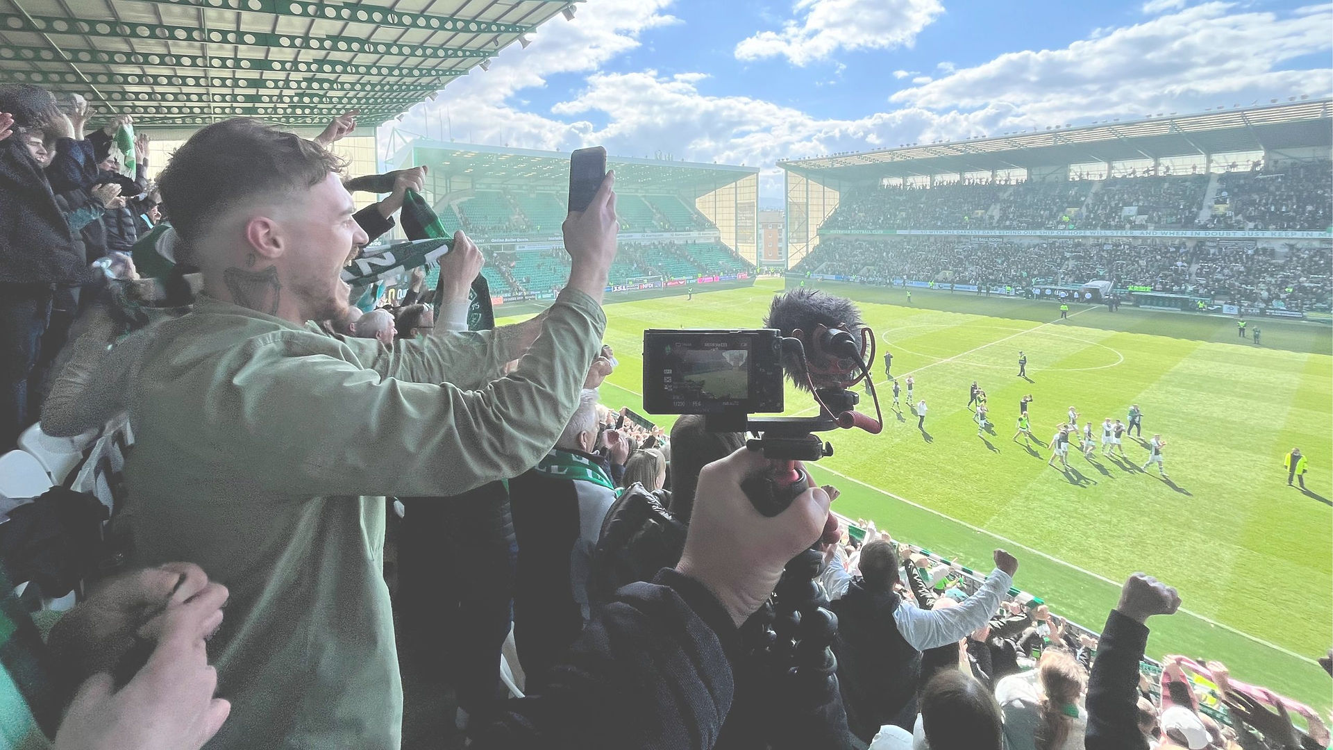 Fotball Stadium, with blue skies and some clouds. Fans cheering, bright pitch with players celebrating.