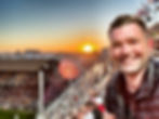 Sports Travel Tom. Man smiling in a stadium at sunset, with crowded seats and flags in the background. The sky is vibrant orange, creating a warm atmosphere.