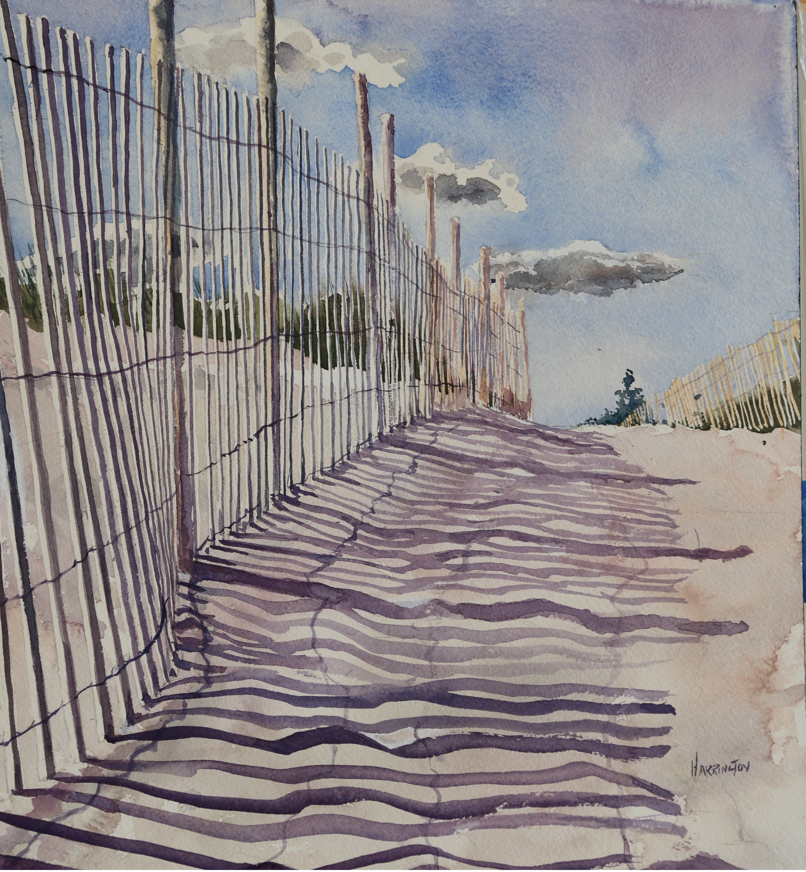 Wooden fence lining the pathway to a beach creates shadows on the uneven sand.