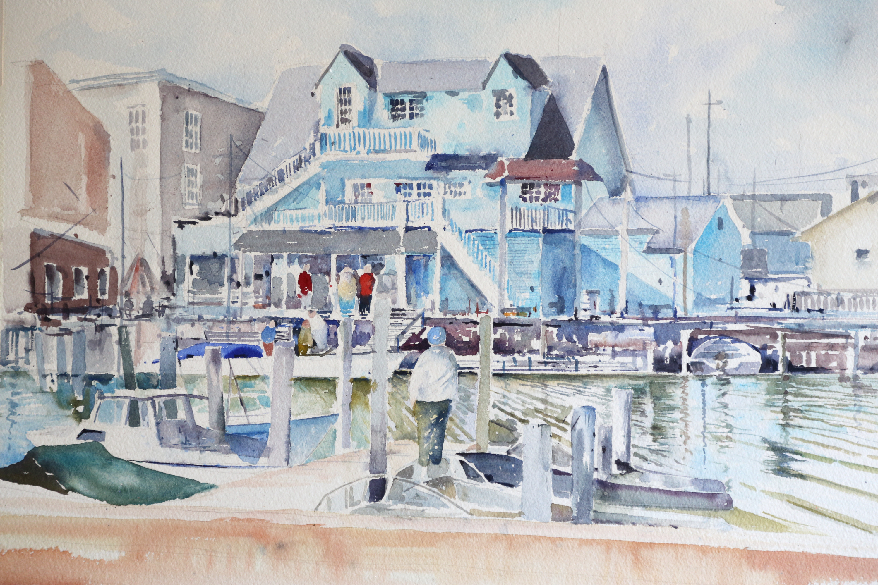 Watercolor painting of waterfront scene: blue building, boats, and figures