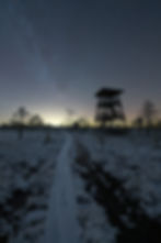 Snowy landscape at night with a wooden watchtower and a star-filled sky. A snow-covered path leads through the quiet, serene scene.