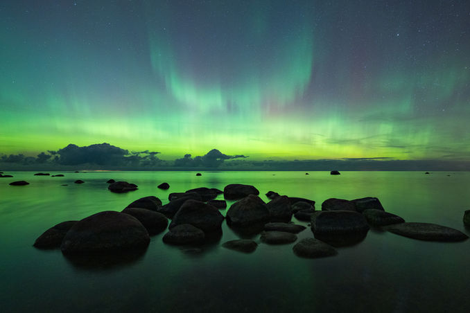 Green auroras dance above a calm seascape, with large rocks scattered across the water's surface. | Rohelised virmalised tantsivad rahuliku meremaastiku kohal, suured kivid on hajutatud üle mere.