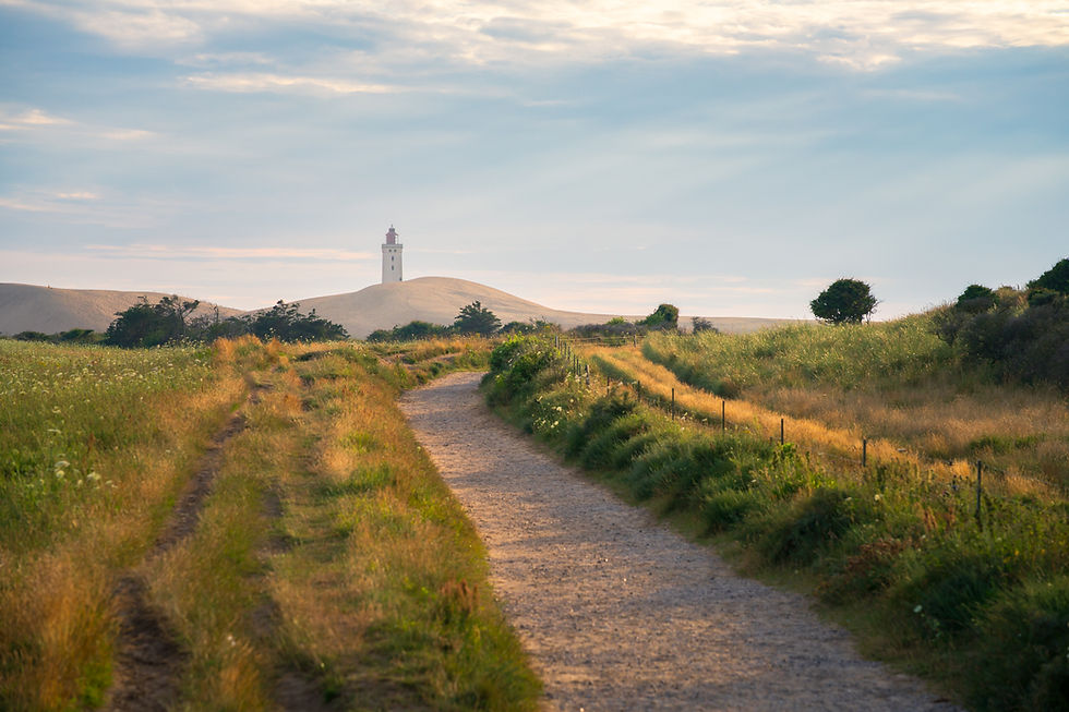 A sandy path leads to a distant lighthouse on a grassy landscape under a partly cloudy sky. Warm light suggests a serene, late afternoon.
