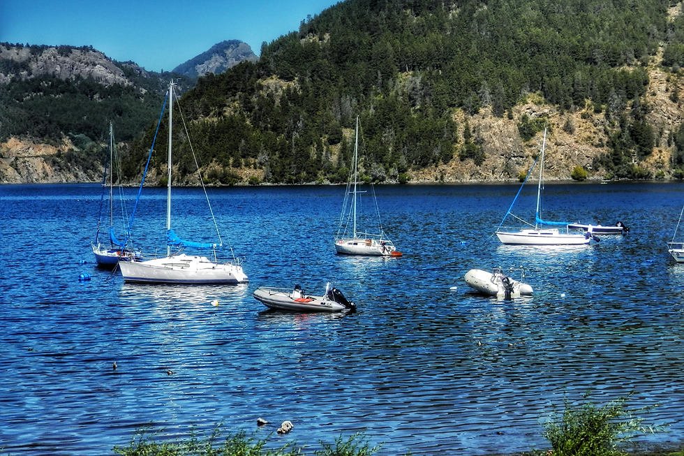 Sailboats and dinghies float on a serene blue lake, surrounded by green hills and distant mountains under a clear sky.