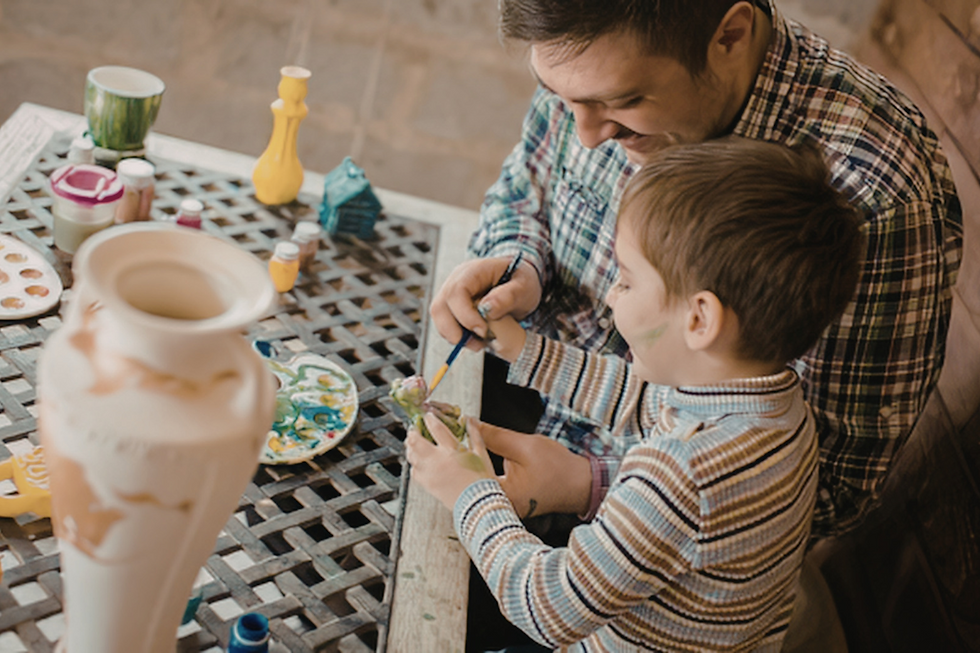 Father and young son paint a ceramic vase together at a table, with paint jars and palettes in a cozy workshop.